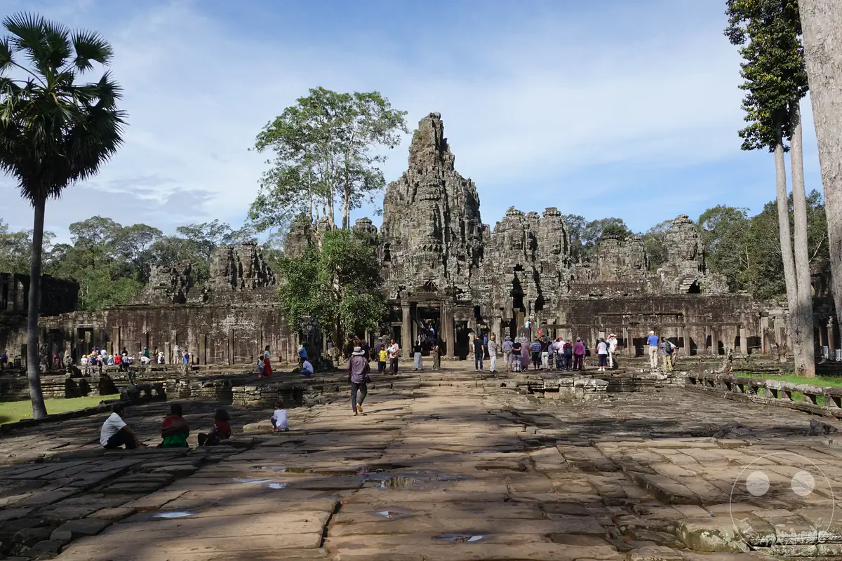 Der historische Bayon-Tempel in Angkor Thom, Kambodscha, mit seinen markanten Steintürmen, umgeben von Bäumen und Besuchern auf dem gepflasterten Vorplatz unter leicht bewölktem Himmel.