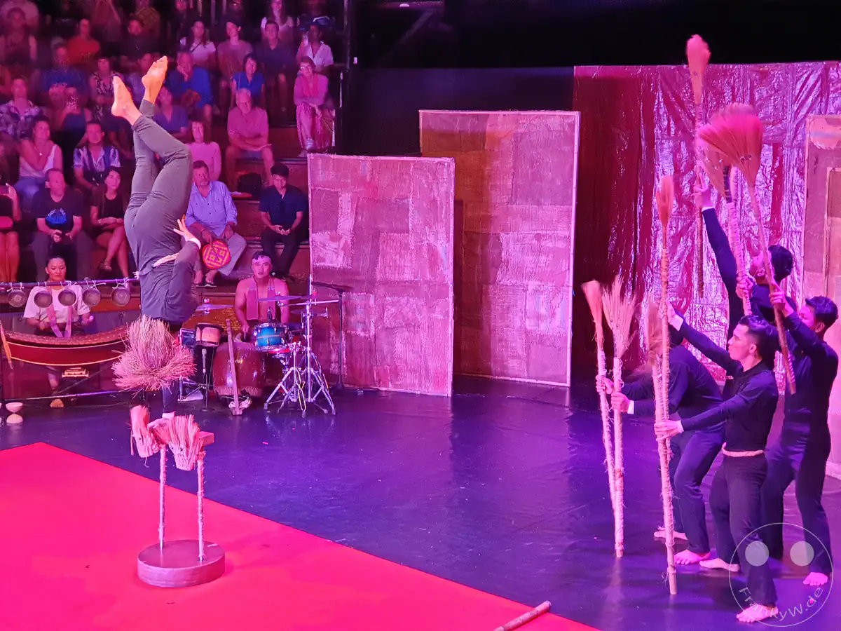 Ein Akrobat balanciert im Handstand auf einem Gestell während einer Live-Aufführung des Phare Kambodschanischen Zirkus in Siem Reap, begleitet von Musikern im Hintergrund und Darstellern mit Strohbesen unter farbiger Bühnenbeleuchtung.