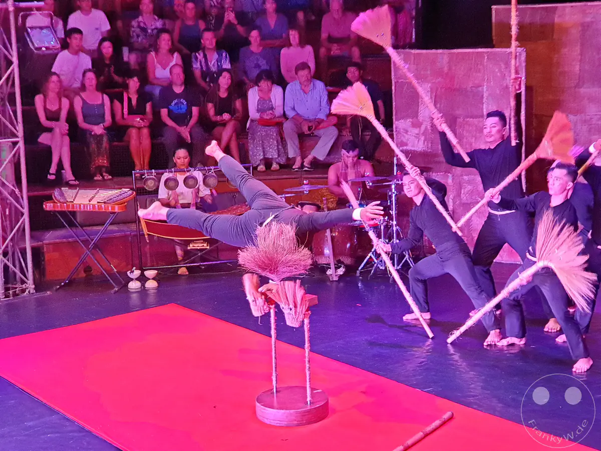 Ein Akrobat balanciert in einer horizontalen Haltefigur auf einem kleinen Handstandgestell, während weitere Darsteller im Hintergrund mit Strohbesen tanzen und Musiker die Live-Show vor Publikum begleiten.