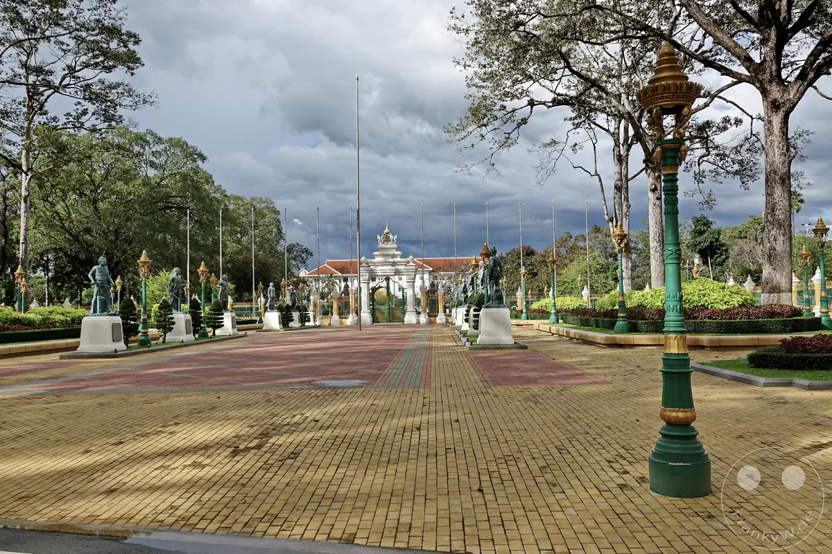 Ein weitläufiger, gepflasterter Vorplatz in den Royal Independence Gardens in Siem Reap mit kunstvoll verzierten, grünen Laternenpfählen, einer Reihe von Statuen auf weißen Sockeln und einem monumentalen Tor im Hintergrund unter bewölktem Himmel.