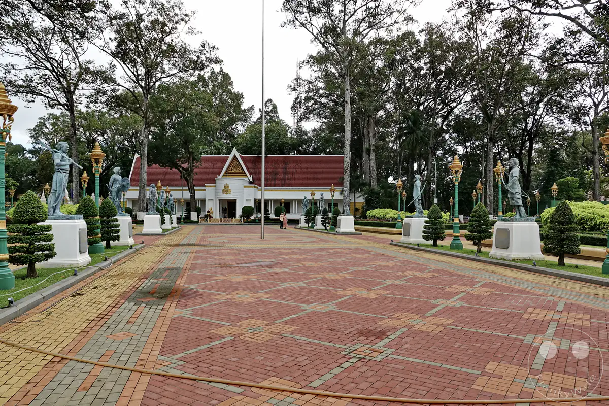 Ein weitläufiger, gemusterter Pflasterweg in den Royal Independence Gardens in Siem Reap, flankiert von historischen Kriegerstatuen auf weißen Sockeln, verzierten Laternenpfählen und hohen Bäumen vor der königlichen Residenz im Hintergrund.