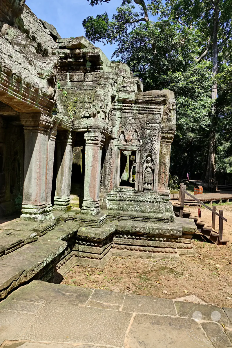 Die historische Außenwand des Ta-Prohm-Tempels in Angkor mit kunstvollen Säulengängen, einem steinernen Fensterrahmen und einem detailreichen Flachrelief einer tanzenden Apsara-Göttin neben einem Holzsteg.