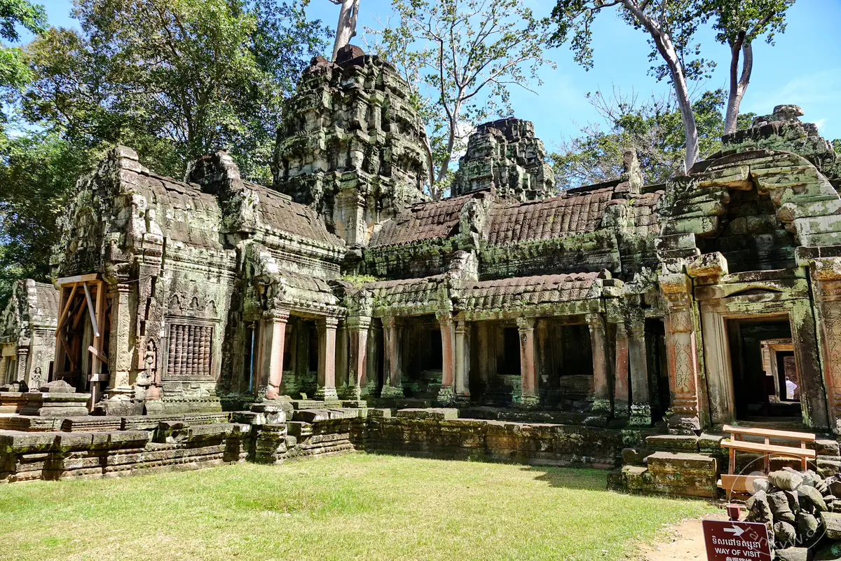 Die weite Außenansicht des historischen Ta-Prohm-Tempels in Angkor mit seinen von Säulen getragenen Galeriebauten und moosbewachsenen Steintürmen vor einer grünen Wiese unter blauem Himmel.