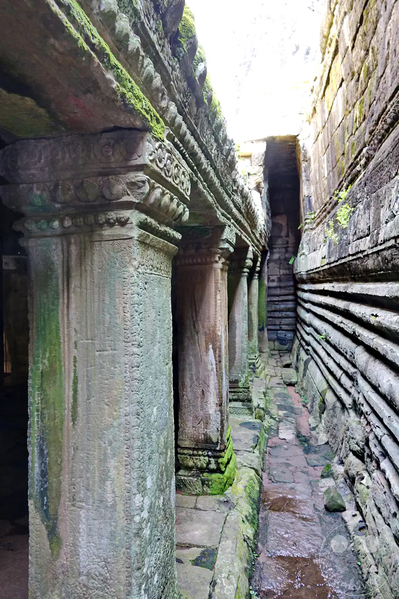 Ein schmaler, historischer Korridor im Ta-Prohm-Tempel in Kambodscha mit verzierten, moosbewachsenen Steinsäulen und einer reliefartigen Außenwand unter einfallendem Tageslicht.