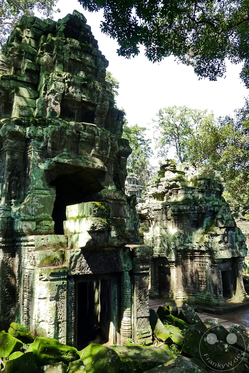 Zwei historische, moosbewachsene Steintürme des Ta-Prohm-Tempels in Angkor inmitten herabgefallener Steinblöcke und dichter Urwaldbäume an einem sonnigen Tag.