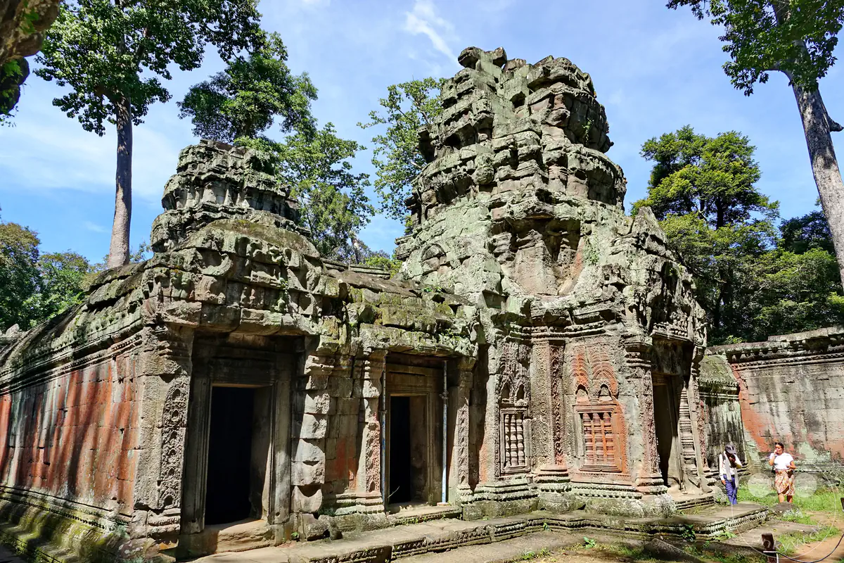 Die historische Außenansicht des Ta-Prohm-Tempels in Angkor mit zwei markanten Steintürmen, detailreichen Giebelreliefs und dunklen Türeingängen vor hohen Dschungelbäumen unter blauem Himmel.