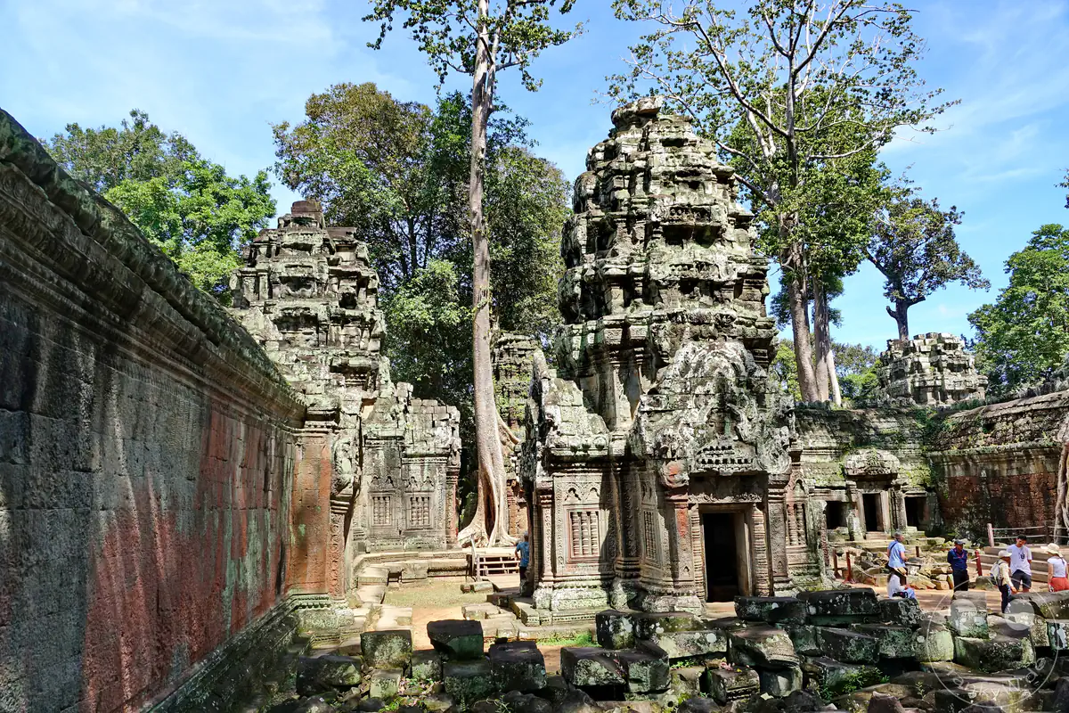 Zwei monumentale, historische Steintürme des Ta-Prohm-Tempels in Angkor mit kunstvollen Schnitzereien inmitten verstreuter Steinblöcke, umgeben von hohen Bäumen und Besuchern unter blauem Himmel.