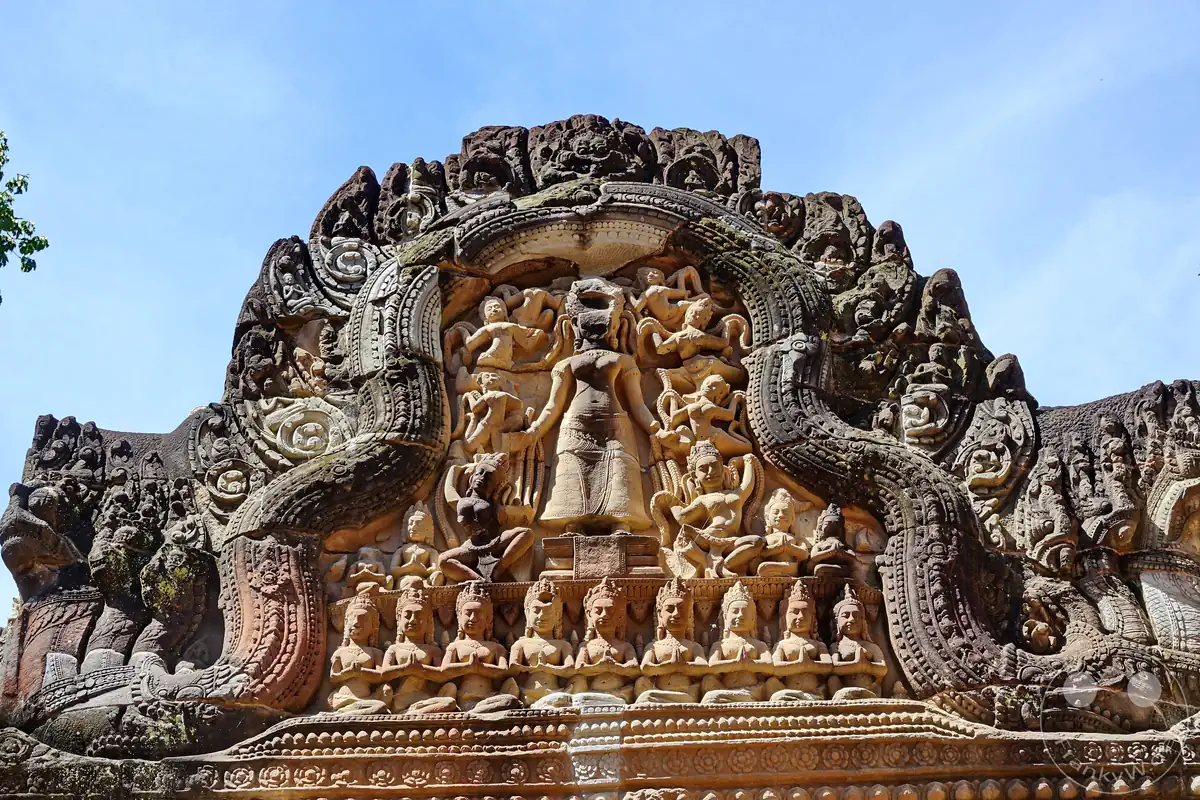 Detailreiches Khmer-Steinrelief auf einem verzierten Sandstein-Giebel des Ta-Prohm-Tempels vor blauem Himmel. Die kunstvolle Gravur zeigt eine zentrale Gottheit, umgeben von tanzenden Apsaras und einer Reihe sitzender Figuren.