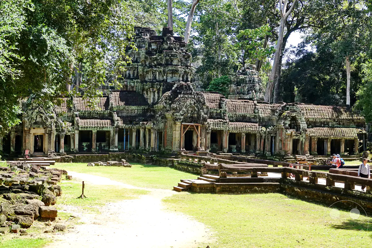 Die weite Außenansicht der historischen, moosbewachsenen Tempelanlage von Ta Prohm in Angkor mit ihren zentralen Steintürmen und weitläufigen Galeriebauten vor einer grünen Rasenfläche und hohen Dschungelbäumen.