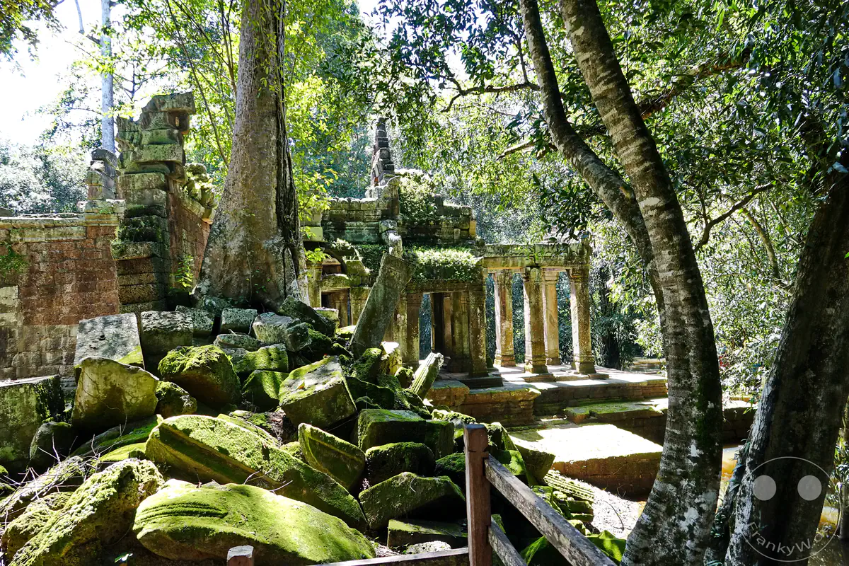 Ein Haufen eingestürzter, moosbewachsener Steinblöcke vor den historischen Säulengängen und Toren des Ta-Prohm-Tempels in Angkor, umgeben von dichten Bäumen im Sonnenlicht.