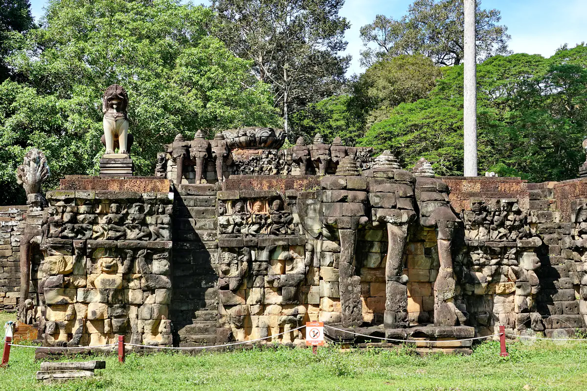 Ein historischer Mauerabschnitt der Terrasse der Elefanten in Angkor Thom mit eingemeißelten Elefantenköpfen, detailreichen Steinreliefs und einer wachenden Löwenstatue auf der oberen Plattform vor dichten Bäumen.