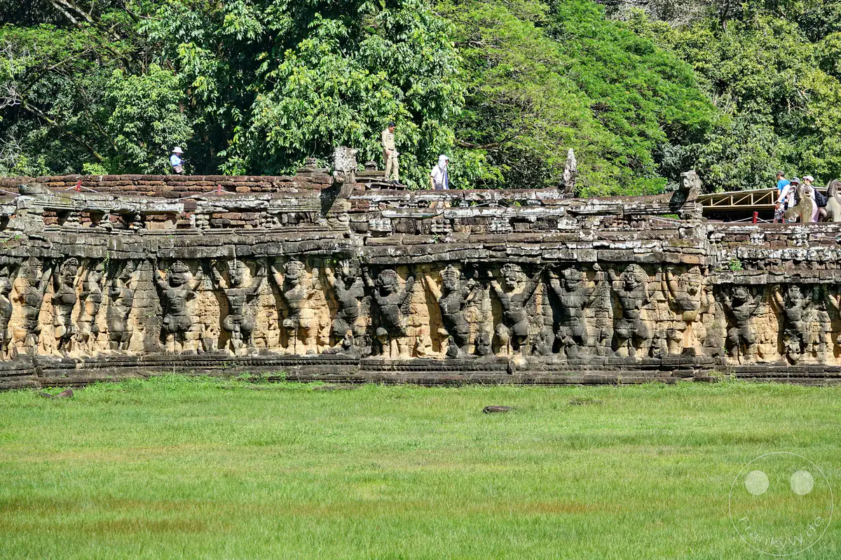 Eine lange Reihe historischer, in den Stein gemeißelter Garuda- und Schutzgott-Reliefs an den Außenmauern der Terrasse des Lepra-Königs in Angkor Thom vor einer weiten, grünen Wiese.