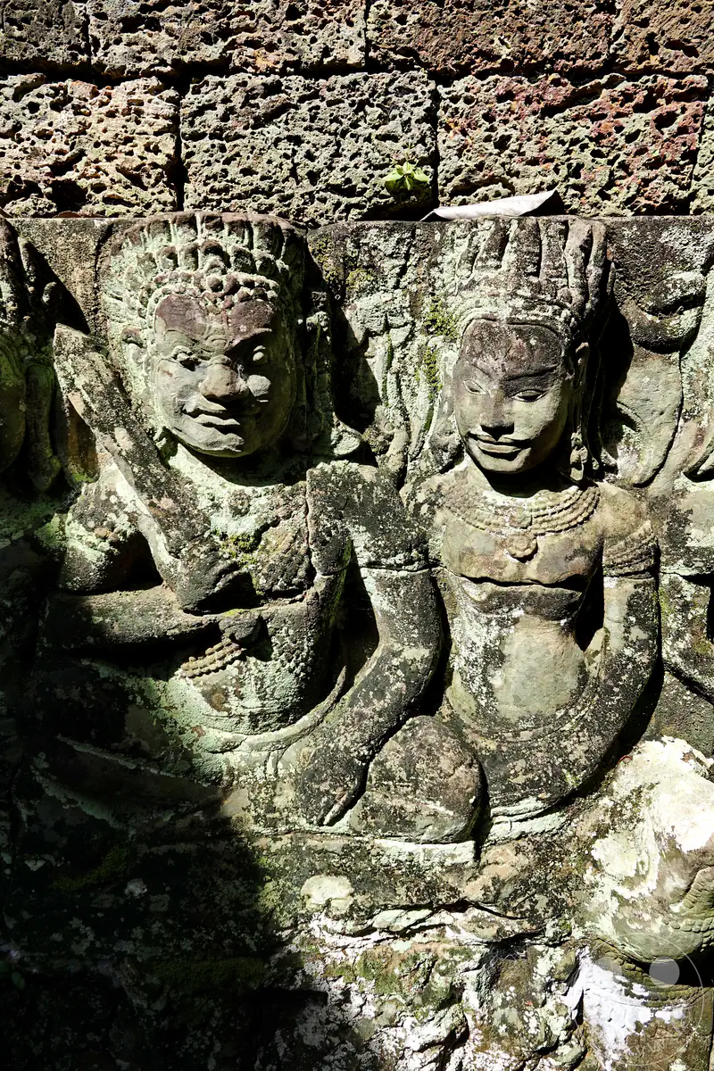 Eine Nahaufnahme von zwei historischen, in den Stein gemeißelten Relief-Skulpturen mit traditionellem Kopfschmuck und Schmuckelementen an der Außenwand der Terrasse des Lepra-Königs in Angkor Thom.
