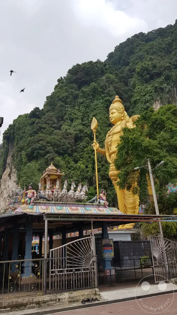 Blick auf die monumentale, goldene Lord-Murugan-Statue am Eingang der Batu Caves bei Kuala Lumpur, Malaysia, mit einem verzierten Tempeldach im Vordergrund und dicht bewachsenen Kalksteinfelsen im Hintergrund.