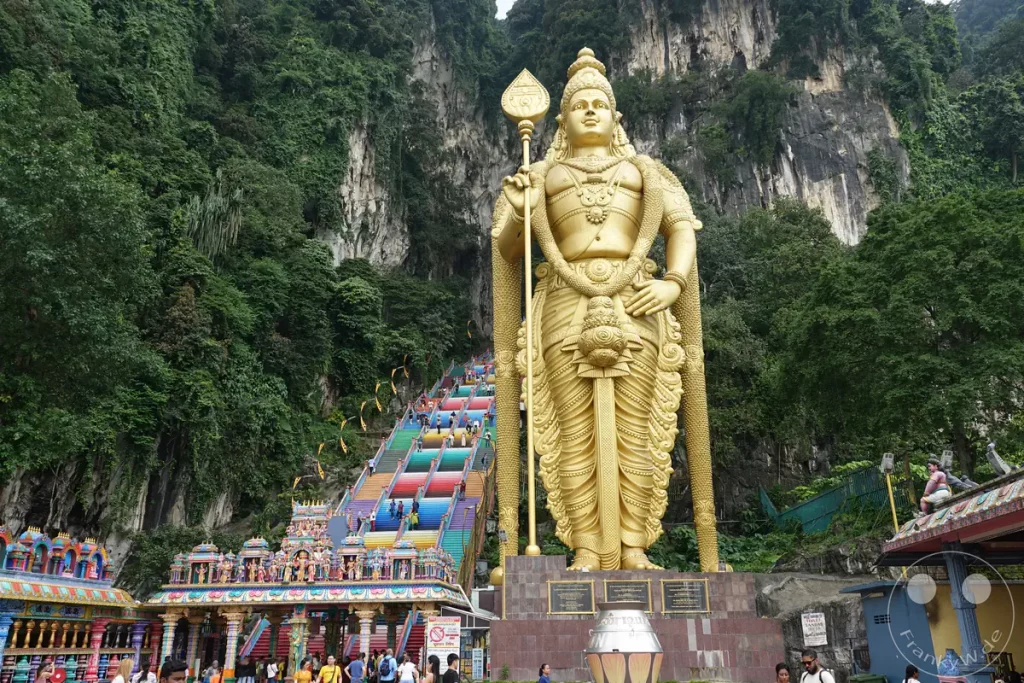 Blick auf die monumentale, goldene Lord-Murugan-Statue und die farbenfrohe Regenbogentreppe am Eingang der Batu Caves bei Kuala Lumpur, Malaysia, umgeben von dicht bewachsenen Kalksteinfelsen und Besuchern auf dem Vorplatz.