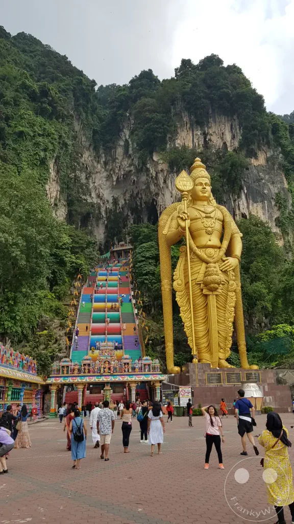 Blick über den gepflasterten Vorplatz auf die gewaltige goldene Lord-Murugan-Statue und die steile, farbenfrohe Regenbogentreppe, die an den bewaldeten Kalksteinfelsen vorbei zum Eingang der Batu Caves bei Kuala Lumpur, Malaysia führt.