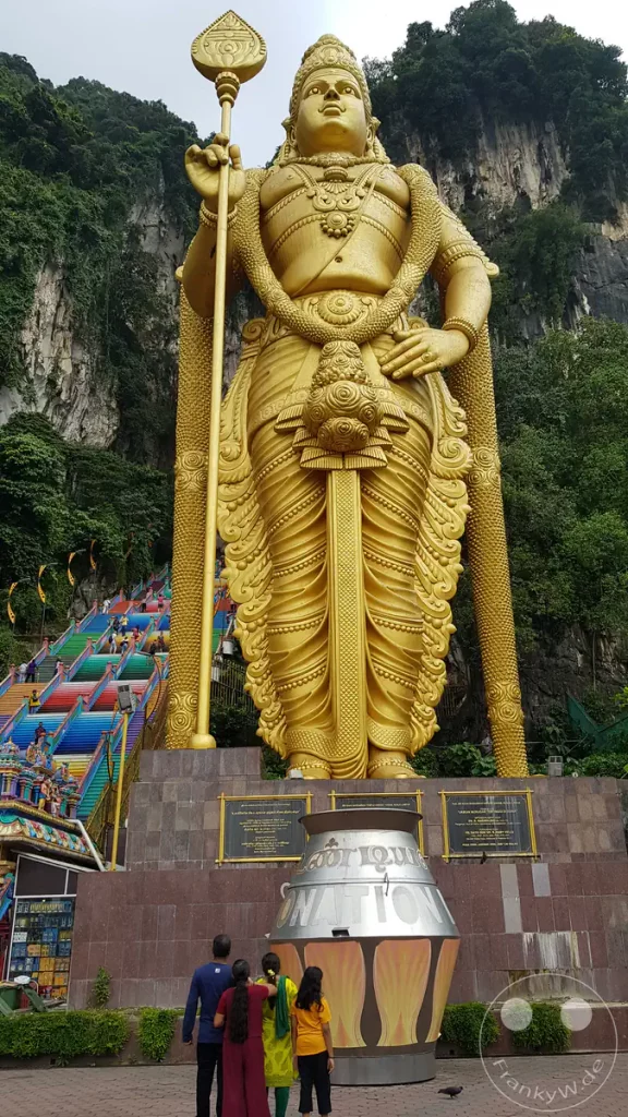 Froschperspektive auf die monumentale, goldene Lord-Murugan-Statue bei den Batu Caves nahe Kuala Lumpur, Malaysia, mit einer großen, krugförmigen Spendenbox im Vordergrund, vor der eine Familie steht, und einem Ausschnitt der farbenfrohen Regenbogentreppe links.