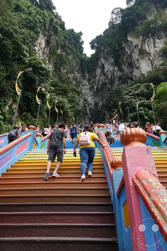 Blick die farbenfrohe Regenbogentreppe hinauf zu den Batu Caves bei Kuala Lumpur, Malaysia, mit zahlreichen Besuchern beim Aufstieg und den steilen, bewaldeten Kalksteinfelsen im Hintergrund.