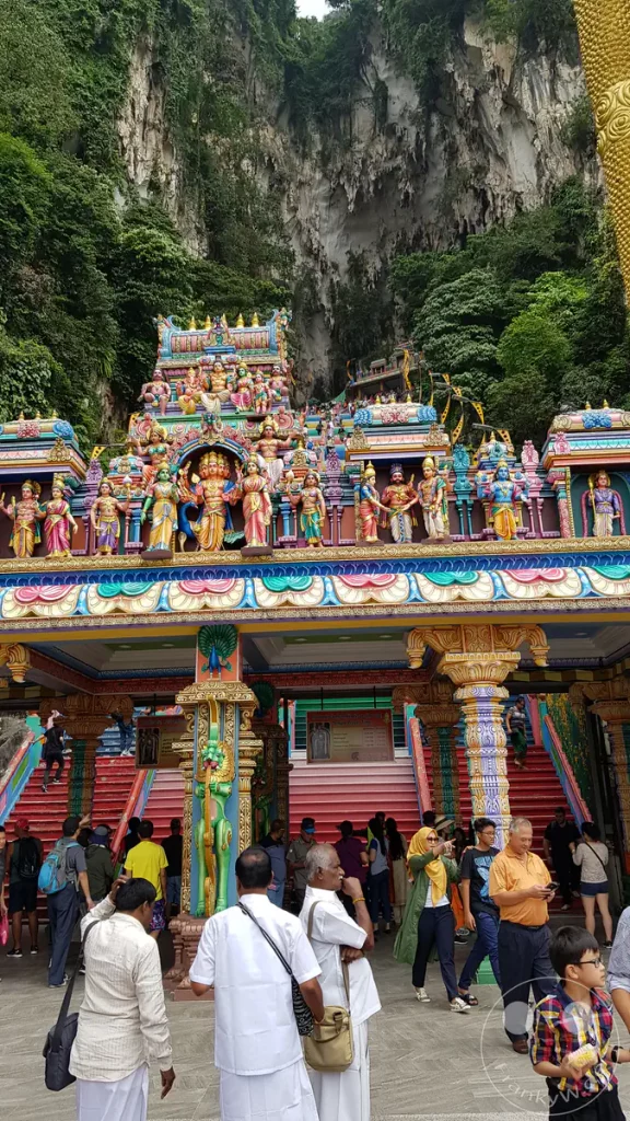 Blick auf den farbenfroh verzierten Eingang und das Gopuram des Sri Subramaniar Swamy Tempels bei den Batu Caves nahe Kuala Lumpur, Malaysia, mit Gläubigen und Besuchern im Vordergrund sowie den steilen Kalksteinfelsen im Hintergrund.