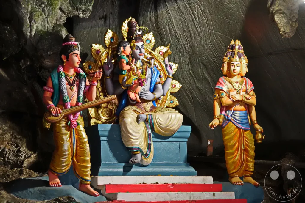 Drei farbenfrohe hinduistische Götterstatuen, darunter Shiva mit einem Kind auf dem Schoß und der vierköpfige Brahma, aufgestellt auf einem kleinen Sockel in einer Nische der Kalksteinhöhle der Batu Caves bei Kuala Lumpur, Malaysia.