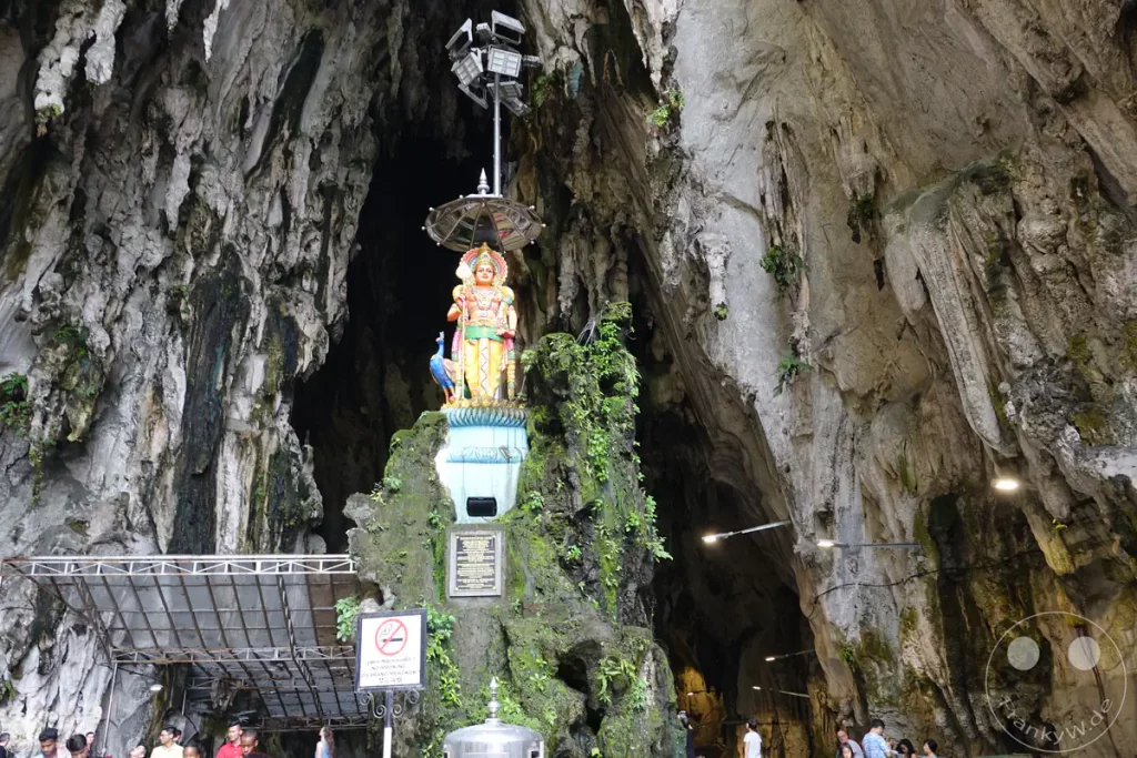 Blick in das Innere der Temple Cave (Kathedralenhöhle) der Batu Caves bei Kuala Lumpur, Malaysia, mit einer erhöhten Lord-Murugan-Statue mitsamt Pfau auf einem bemoosten Felsen inmitten einer gewaltigen Kalksteinhöhle mit herabhängenden Stalaktiten.