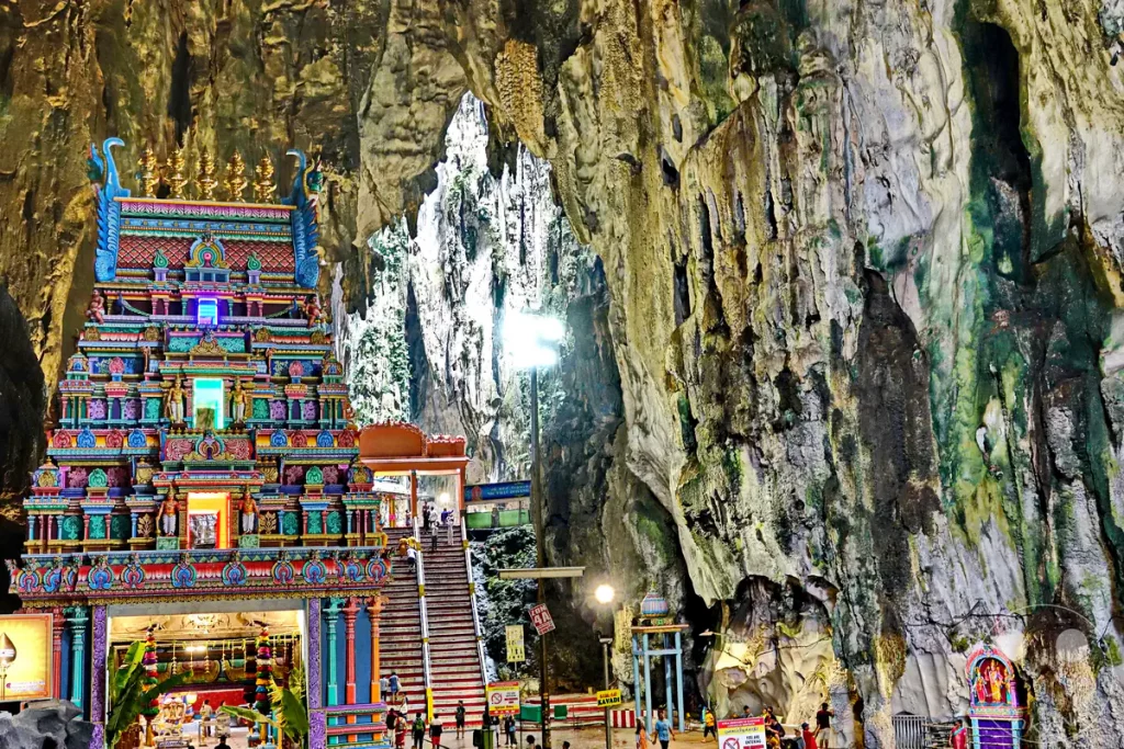Blick in das Innere der gewaltigen Temple Cave der Batu Caves bei Kuala Lumpur, Malaysia, mit dem farbenprächtig verzierten, mehrstöckigen Gopuram eines Hindu-Tempels vor einer steilen Treppe und den monumentalen, naturbelassenen Wänden der Kalksteinhöhle.