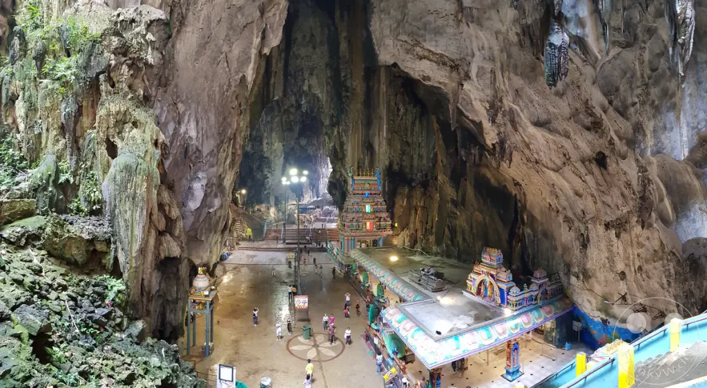 Panoramablick aus erhöhter Perspektive in das Innere der gewaltigen Temple Cave der Batu Caves bei Kuala Lumpur, Malaysia, mit dem bunt verzierten Sri Valli Devanayagar Murugan Tempel, kleinen Besucher- und Pilgergruppen auf dem Höhlenboden und monumentalen, zerklüfteten Kalksteinwänden.