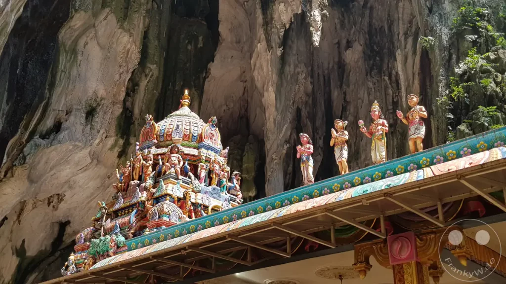 Blick auf ein kunstvoll verziertes, buntes Tempeldach mit einer Kuppel und mehreren stehenden Götterstatuen im Inneren der monumentalen Kalksteinhöhle der Batu Caves bei Kuala Lumpur, Malaysia.