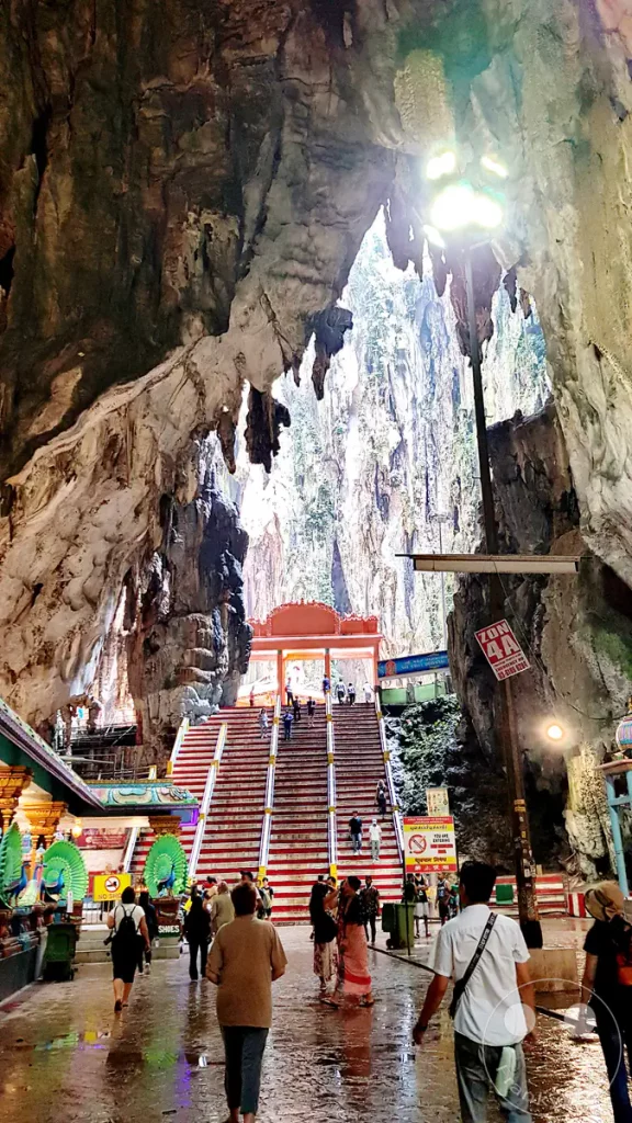 Blick aus dem Inneren der gewaltigen Temple Cave der Batu Caves bei Kuala Lumpur, Malaysia, in Richtung einer rot-weißen Treppe, die zu einem weiteren Höhlenabschnitt führt, umrahmt von monumentalen Kalksteinformationen und herabhängenden Stalaktiten sowie Besuchern im Vordergrund.