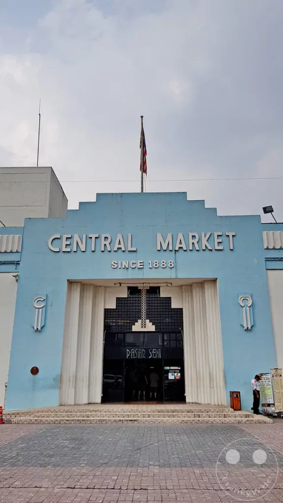 Der hellblaue Art-Deco-Haupteingang des Central Market (Pasar Seni) in Kuala Lumpur, Malaysia, mit dem Schriftzug „CENTRAL MARKET SINCE 1888“, einer malaysischen Flagge auf dem Dach und geometrischen Säulenelementen am Portal.