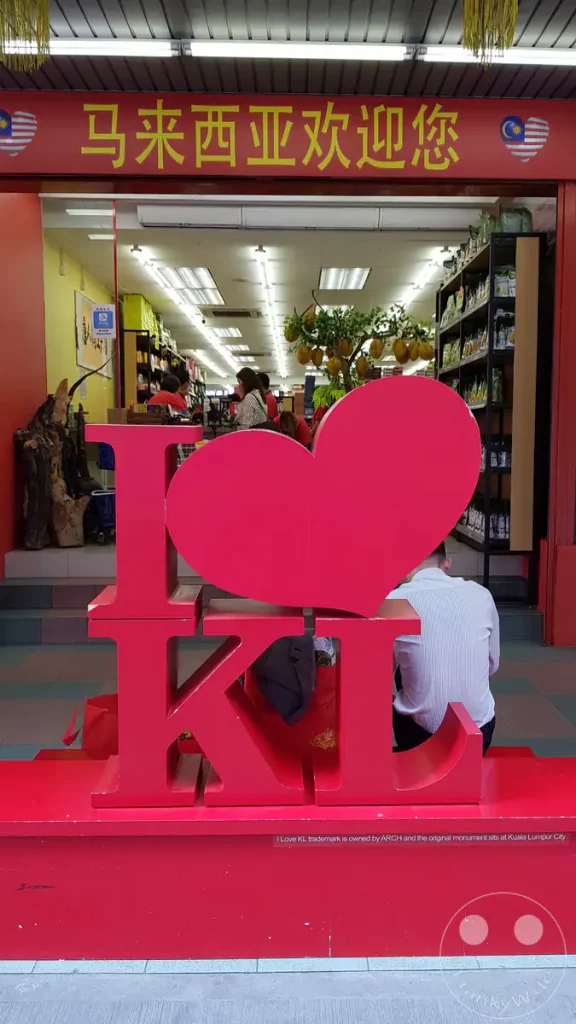 Nahaufnahme einer großen roten „I Love KL“-Skulptur auf einem roten Sockel vor dem Eingang eines hell erleuchteten Souvenirgeschäfts mit chinesischen Schriftzeichen und malaysischen Flaggenmotiven in Kuala Lumpur, Malaysia.