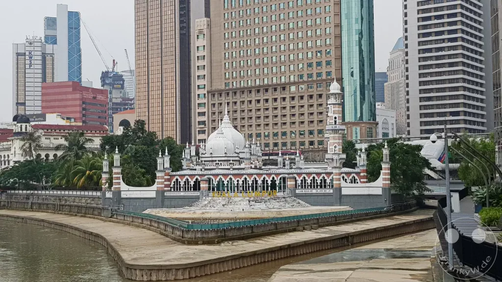 Die historische, weiße Jamek-Moschee mit ihren markanten Minaretten und Kuppeln an der Landspitze des „River of Life“ in Kuala Lumpur, Malaysia, umgeben vom Zusammenfluss der Flüsse Klang und Gombak sowie modernen Hochhäusern im Hintergrund.