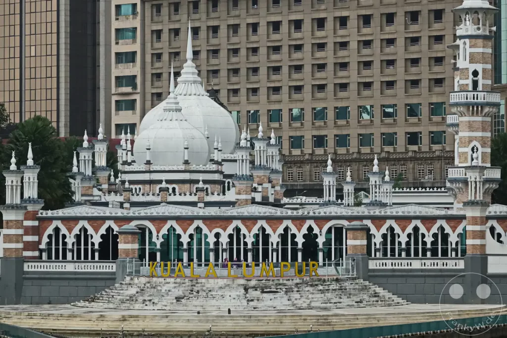 Die detailreiche Fassade der historischen Jamek-Moschee in Kuala Lumpur, Malaysia, mit ihren weißen Kuppeln, filigranen Minaretten und maurischen Bögen am Ufer des River of Life, vor einem großen modernen Bürogebäude im Hintergrund.