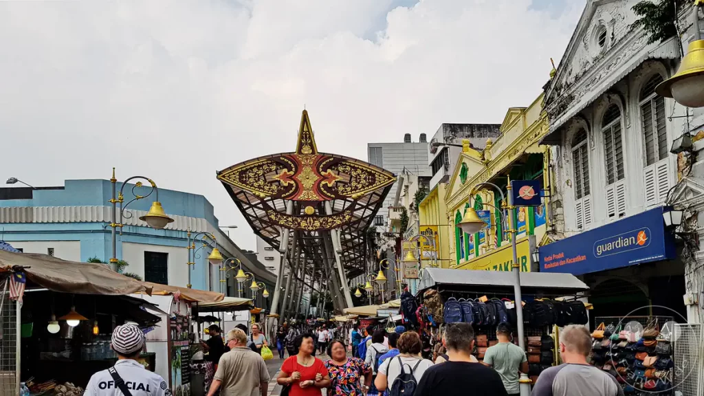 Blick entlang des belebten Kasturi Walk neben dem Central Market in Kuala Lumpur, Malaysia, mit einer monumentalen Überdachung in Form eines traditionellen malaysischen Drachens (Wau Bulan), historischen Shophouses mit einer Guardian-Filiale auf der rechten Seite und zahlreichen Besuchern.