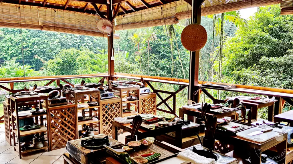 Ein überdachter, offener Holzpavillon der Lazat Cooking School in Kuala Lumpur, Malaysia, eingerichtet für einen Kochkurs mit mehreren hölzernen Arbeitsstationen, tragbaren Gaskochern, Schneidebrettern und vorbereiteten Zutaten vor einer Kulisse aus dichtem, grünem Regenwald.