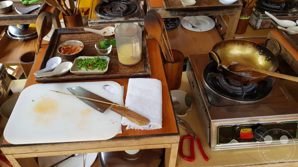 Eine hölzerne Arbeitsstation bei einem malaysischen Kochkurs in der Lazat Cooking School in Kuala Lumpur, Malaysia, mit einem Gaskocher der Marke Butterfly, einer goldfarbenen Pfanne, einem Schneidebrett mit Küchenmesser, Kochutensilien aus Holz sowie vorbereiteten Zutaten wie geschnittenen Chilis und Gewürzen auf kleinen Tellern.