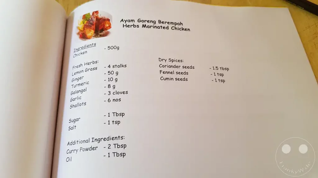Eine geöffnete Seite eines Rezeptbuchs mit der Zutatenliste für „Ayam Goreng Berempah“ (Herbs Marinated Chicken), gedruckt in schwarzer Schrift auf weißem Papier mit einem kleinen Produktbild oben links.
