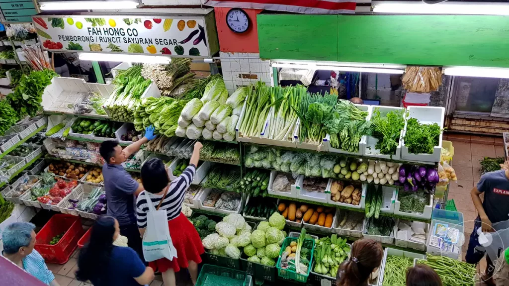 Ein reich bestückter Gemüsestand der Firma Fah Hong Co auf dem Pasar Besar Taman Tun Dr. Ismail (TTDI Market) in Kuala Lumpur, Malaysia, an dem Kunden frisches asiatisches Gemüse wie Chinakohl, Frühlingszwiebeln, Pak Choi, Auberginen und Gurken auswählen, die in weißen Styroporkisten und grünen Körben ordentlich gestapelt sind.