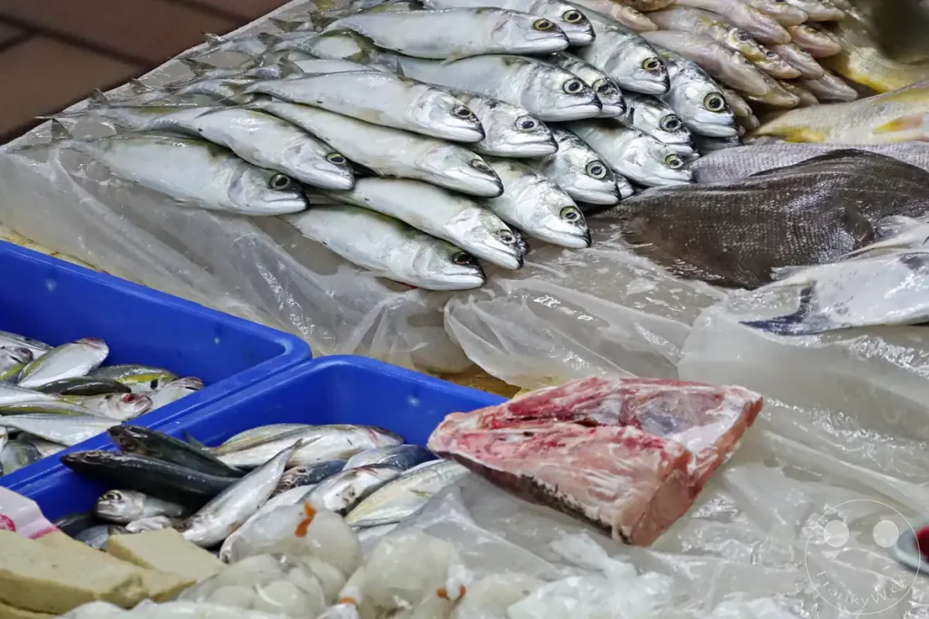 Frischer Fisch, darunter ordentlich aufgereihte Makrelen auf einer eisgekühlten Plastikfolie, kleinere Fische in blauen Kunststoffkisten und ein portioniertes Fischstück im Vordergrund an einem Marktstand auf dem Pasar Besar Taman Tun Dr. Ismail in Kuala Lumpur, Malaysia.