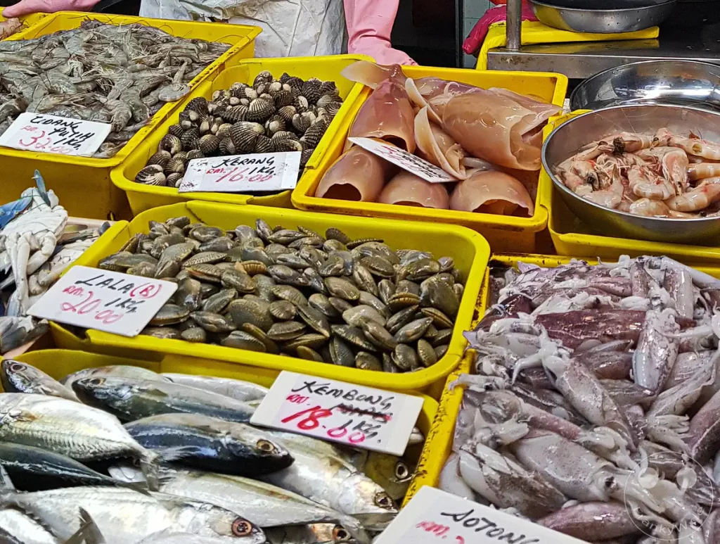 Frische Meeresfrüchte und Speisefische in gelben Kunststoffkisten an einem Marktstand auf dem Pasar Besar Taman Tun Dr. Ismail in Kuala Lumpur, Malaysia, darunter Kalmare, verschiedene Muschelsorten wie Herzmuscheln (Kerang) und Teppichmuscheln (Lala), Garnelen sowie Makrelen (Kembong) mit handschriftlichen Preisschildern.