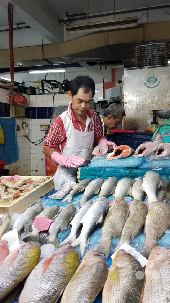 Ein Fischverkäufer mit Schürze und pinkfarbenen Gummihandschuhen an seinem Marktstand auf dem Pasar Besar Taman Tun Dr. Ismail in Kuala Lumpur, Malaysia, hinter einer großen Auslage mit frischem Fisch wie Lachs, Zackenbarsch und Barramundi auf zerstoßenem Eis.