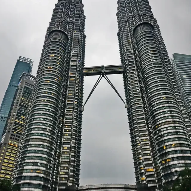 Die symmetrisch aufragenden Petronas Twin Towers in Kuala Lumpur, Malaysia, mit ihrer charakteristischen Skybridge, dem beleuchteten Eingangsbereich des Suria-KLCC-Einkaufszentrums am Fuß der Gebäude und zahlreichen Besuchern im Vordergrund unter einem grauen Himmel.