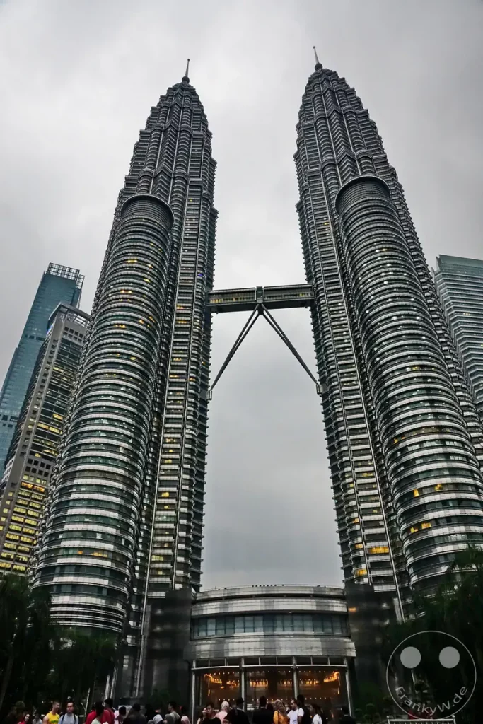 Die symmetrisch aufragenden Petronas Twin Towers in Kuala Lumpur, Malaysia, mit ihrer charakteristischen Skybridge, dem beleuchteten Eingangsbereich des Suria-KLCC-Einkaufszentrums am Fuß der Gebäude und zahlreichen Besuchern im Vordergrund unter einem grauen Himmel.