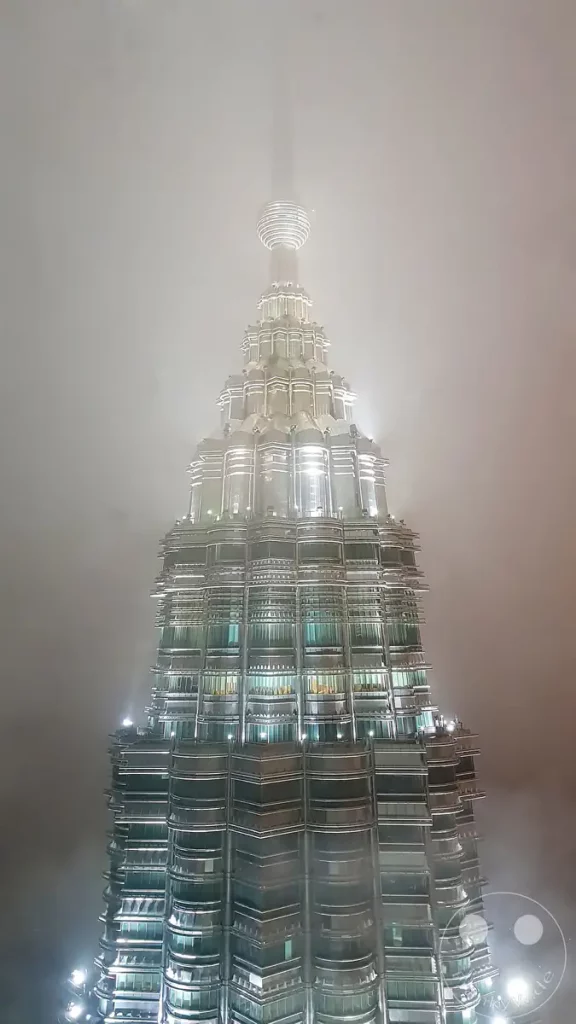Die hell erleuchtete, stufenförmige Turmspitze eines der Petronas Twin Towers in Kuala Lumpur, Malaysia, während einer Nachtaufnahme, bei der die oberste Nadel im dichten Nebel und Dunst verschwindet.
