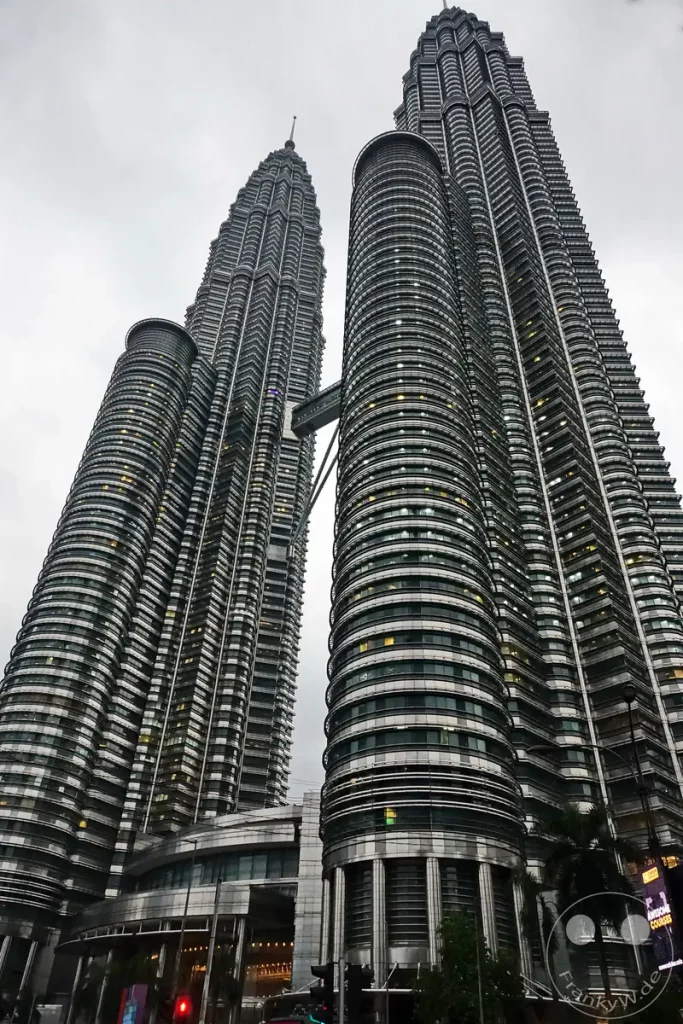 Die hoch aufragenden Petronas Twin Towers in Kuala Lumpur, Malaysia, mit ihrer markanten, metallisch glänzenden Glasfassade, den abgestuften Turmspitzen und der verbindenden Skybridge im oberen Drittel unter einem wolkenverhangenen Himmel.