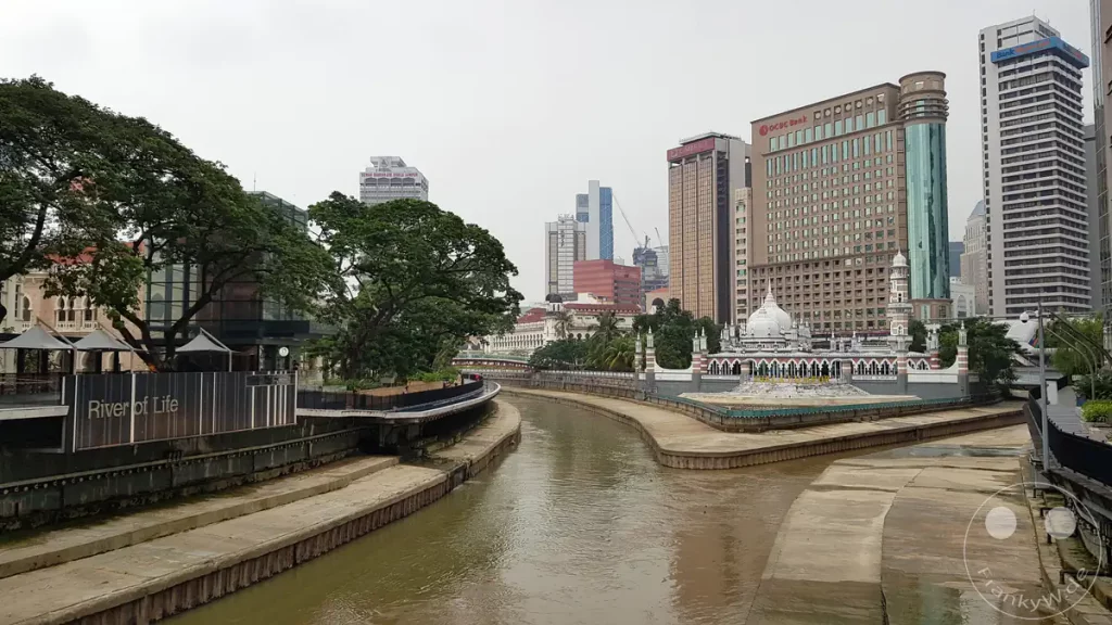 Der Zusammenfluss der Flüsse Klang und Gombak am „River of Life“ in Kuala Lumpur, Malaysia, mit der historischen, weißen Jamek-Moschee an der Landspitze, modernen Bürohochhäusern wie dem OCBC-Bank-Gebäude im Hintergrund und einer Uferpromenade mit großen, grünen Bäumen auf der linken Seite.