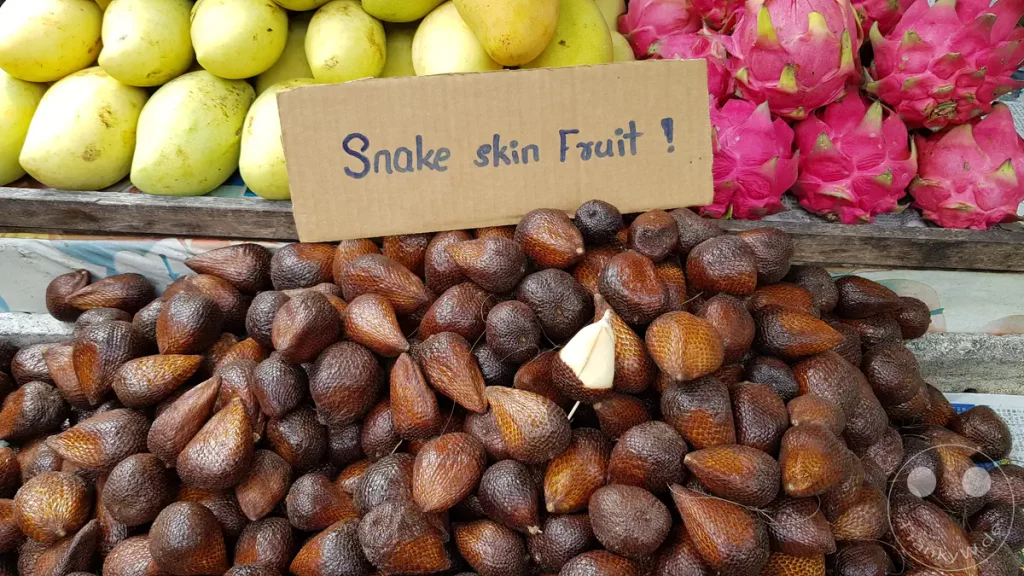 Draufsicht auf einen Marktstand in Kuala Lumpur, Malaysia, mit einem großen Haufen brauner, geschuppter Schlangenfrüchte (Salak), einem handschriftlichen Pappschild mit der Aufschrift „Snake skin Fruit!“, sowie gelben Mangos und pinkfarbenen Drachenfrüchten im Hintergrund.