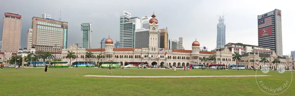 Ein breites Panorama des historischen Sultan Abdul Samad Buildings mit seinen charakteristischen Kuppeln und dem zentralen Uhrturm am Dataran Merdeka in Kuala Lumpur, Malaysia, umgeben von der weitläufigen grünen Rasenfläche im Vordergrund sowie modernen Wolkenkratzern wie dem im Bau befindlichen Merdeka 118 und dem HSBC-Gebäude im Hintergrund.