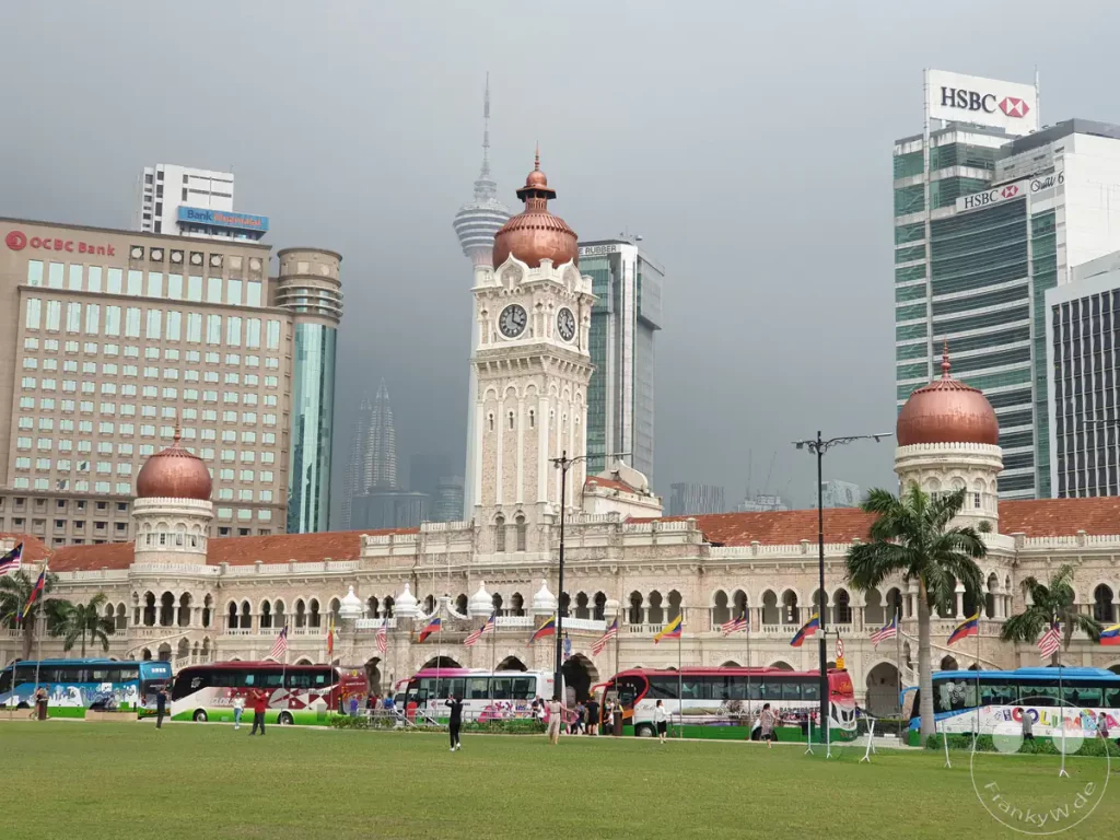 Das historische Sultan Abdul Samad Building mit seinem markanten, kupfergedeckten Uhrturm und kleineren Kuppeln am Rande der Rasenfläche des Dataran Merdeka in Kuala Lumpur, Malaysia, mit Reisebussen im Vordergrund sowie dem KL Tower, den Spitzen der Petronas Towers und modernen Bankgebäuden im nebligen Hintergrund.