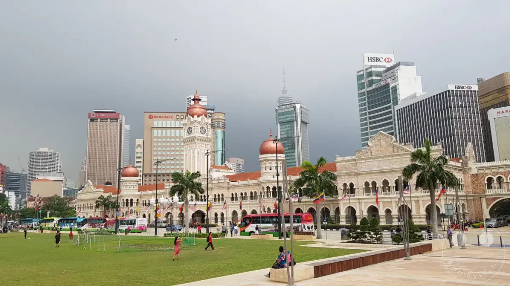 Das historische Sultan Abdul Samad Building mit seinen kupferfarbenen Kuppeln und dem markanten Uhrturm am Rande des grünen Dataran Merdeka (Unabhängigkeitsplatz) in Kuala Lumpur, Malaysia, umgeben von Palmen, vorbeifahrenden Reisebussen und modernen Wolkenkratzern wie den HSBC- und CIMB-Bankgebäuden im Hintergrund unter einem bewölkten Himmel.